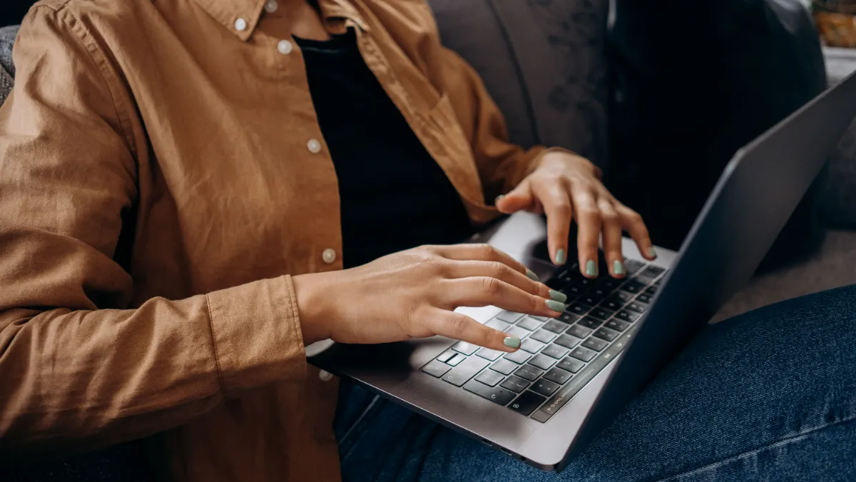 A person in a tan jacket typing on a laptop balanced on their knees